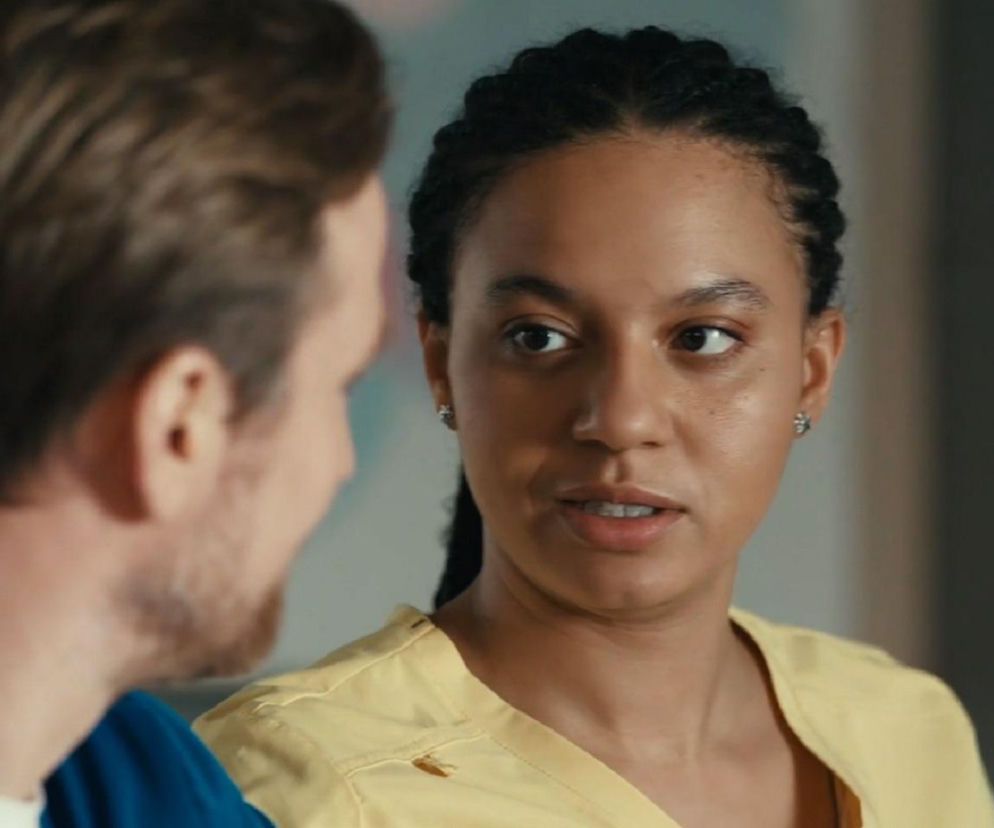 Maria (Sylwia Achu) w żółtym uniformie lekarskim, ze zmartwioną miną, patrzy na Michała (Mateusz Janicki), który stoi do niej odwrócony plecami. Kobieta ma splątane włosy i kolczyki w uszach. To kadr z serialu Na dobre i na złe, o którym przeczytasz więcej na Super Seriale.