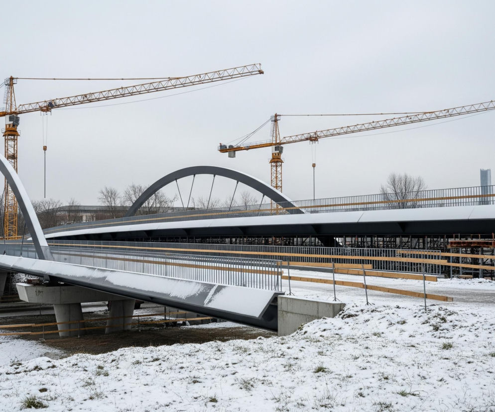Na pierwszym planie, rozległy teren pokryty jest warstwą śniegu, odsłaniając sporadyczne kępki suchej trawy. Centralną część obrazu zajmuje niedokończony, nowoczesny most z dwiema stalowymi, szarymi, łukowatymi konstrukcjami, z których zwisają czarne liny podtrzymujące płaskie jezdnie, również pokryte śniegiem. Na drugim planie dominują dwie duże, żółte żurawie budowlane z długimi ramionami, pracujące nad dalszą częścią mostu i jego betonowymi, szarymi filarami, otoczonymi drewnianymi deskami szalunkowymi. Tło wypełnia szare, pochmurne niebo, a horyzont zamykają zarysy drzew bez liści i odległych budynków.