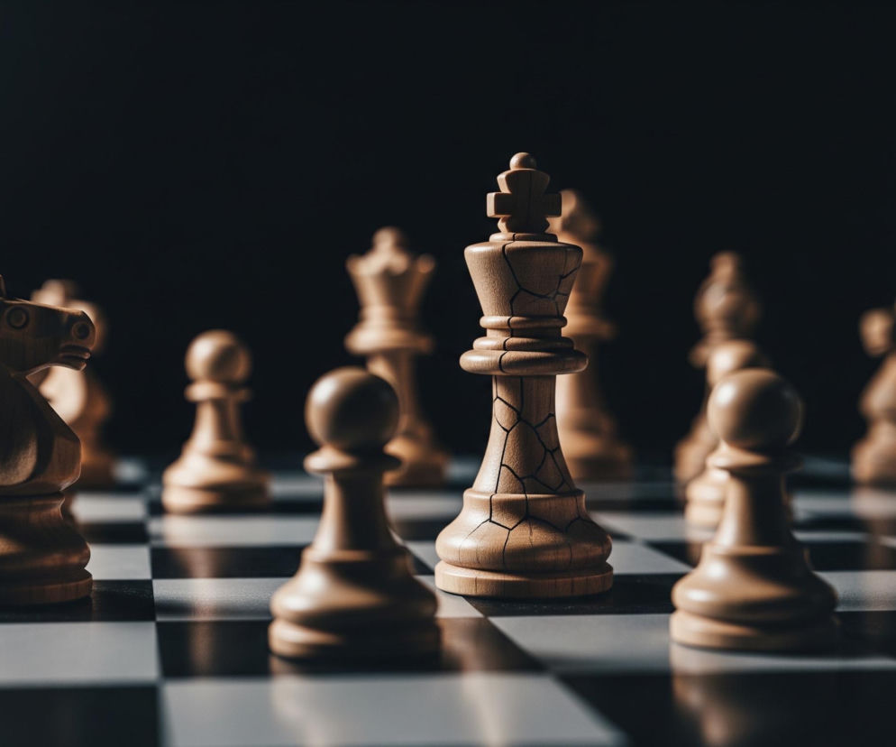 Częściowo oświetlona drewniana szachownica z figurami. Głównym, najbardziej widocznym obiektem jest jasny król stojący w centrum, z widocznymi pęknięciami na korpusie, co nadaje mu postarzony wygląd. Po jego lewej stronie, bliżej krawędzi obrazu, stoi jasny skoczek, a przed nim, na pierwszym planie, jasny pionek. Większość figur jest jasnego koloru drewna, a tło jest ciemne i rozmyte, co skupia uwagę na figurach znajdujących się na szachownicy.