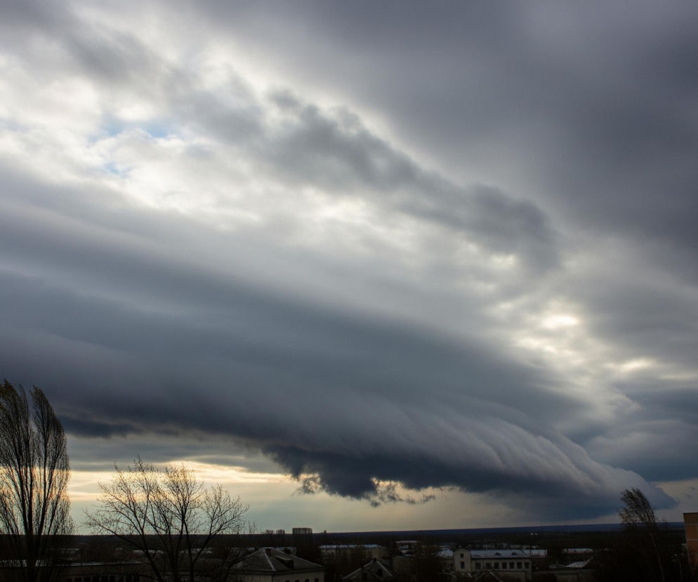 Ponad miastem rozciąga się rozległe, ciemne zachmurzenie, przy czym duża, szara chmura w kształcie fali, z wyraźnymi poziomymi pasami, dominuje środek kadru. Od prawej strony dołem chmury, a także na górze po prawej, przebijają się jaśniejsze fragmenty nieba, oświetlone światłem słonecznym. Horyzont na dole obrazu ukazuje linię niskich budynków mieszkalnych o stonowanych kolorach, z ciemnymi konturami drzew i krzewów na pierwszym planie, oraz niewielkimi fragmentami błękitnego nieba widocznymi między chmurami w lewej górnej części.