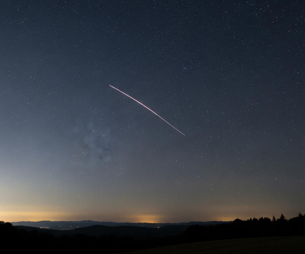 Rozgwieżdżone niebo dominujące na zdjęciu przechodzi od ciemnego granatu u góry w jaśniejszy, błękitno-szary odcień w kierunku horyzontu. W poprzek nieba biegnie długi, jasnoróżowy smug światła, prawdopodobnie ślad meteoru. Poniżej rozległego nieba widoczna jest ciemna sylwetka zalesionego wzgórza, z pojedynczymi, ostrymi konturami drzew na tle delikatnej pomarańczowej łuny miejskiego światła, widocznej nad odległymi pagórkami.