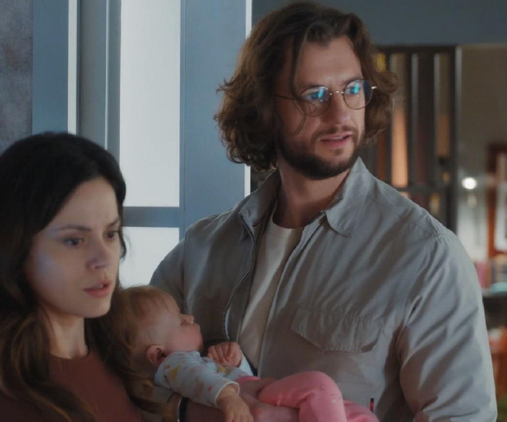 Mariusz (Mateusz Mosiewicz) trzymający śpiącą córkę Zosię, zaniepokojony patrzy na Kasię (Paulina Lasota), która stoi obok z zatroskaną miną. Scena z serialu M jak miłość, gdzie Mariusz przerwie rozmowę Kasi i Anety o dziecku. Więcej informacji na Super Seriale.