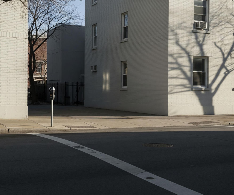 Narożnik ulicy z budynkami po obu stronach i chodnikami. Czarne, asfaltowe jezdnie z białą linią malowaną na nawierzchni i okrągłymi studzienkami kanalizacyjnymi, przecinają się na pierwszym planie. Po lewej stronie widać parkingowy słupek pomiarowy i metalowy płot, za którym widać ciemnoszary budynek, a za nim fragment ceglanego budynku z nagimi drzewami. Po prawej, jasny ceglany budynek z licznymi oknami i jednostką klimatyzacyjną na piętrze, rzuca długie cienie gałęzi drzew na swoją ścianę.