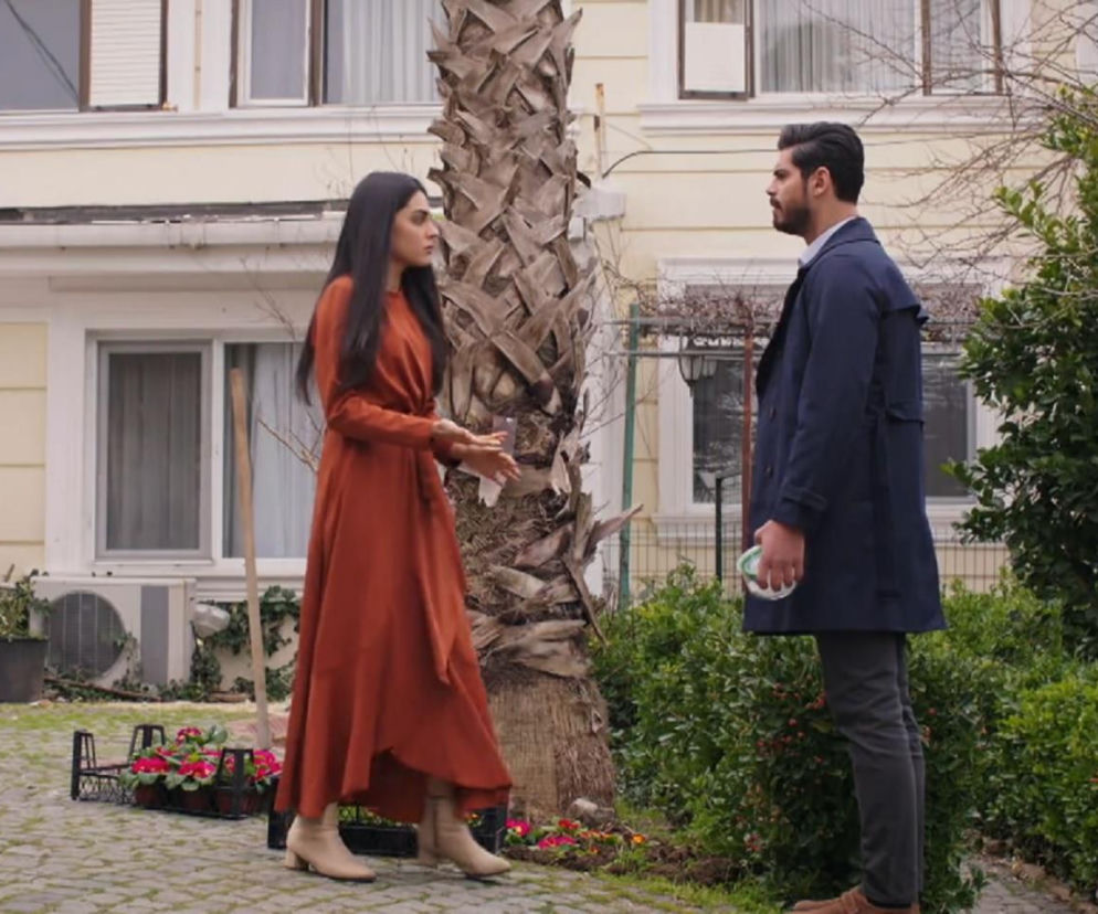 Hancer i Cihan rozmawiają w ogrodzie, stojąc naprzeciwko siebie obok palmy. Kobieta w rudej sukience i beżowych butach gestykuluje, mężczyzna w granatowym płaszczu i ciemnych spodniach słucha. Za nimi widać jasny dom. Więcej o ich losach na Super Seriale.