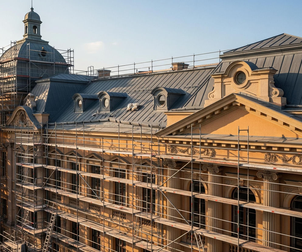 Duży budynek o piaskowej elewacji, pokryty w większości rusztowaniami. Centralną częścią obiektu jest kopuła z iglicą, otoczona metalowymi elementami konstrukcji wsporczej, a obok niej rozciąga się ciemnoszary dach z licznymi lukarnami o okrągłych oknach. Na parterze i piętrze budynku, między rusztowaniami, widać rzędy prostokątnych okien oraz ozdobne pilastry i gzymsy. W tle, po lewej stronie, zza budowy wyłaniają się zarysy innych budynków oraz fragmenty infrastruktury, prawdopodobnie torów kolejowych lub drogi.