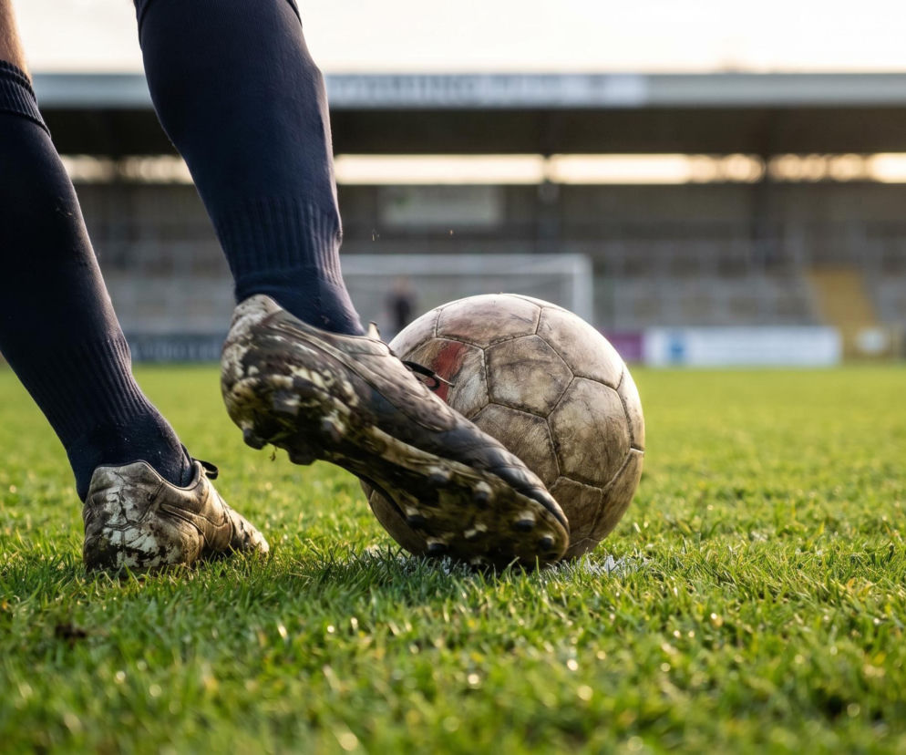 Piłkarz w ciemnych skarpetach i ubłoconych korkach uderza starą piłkę na tle murawy stadionu, symbolizując historyczne osiągnięcie Harry'ego Kane'a na mistrzostwach świata, o którym przeczytasz na naszym portalu.
