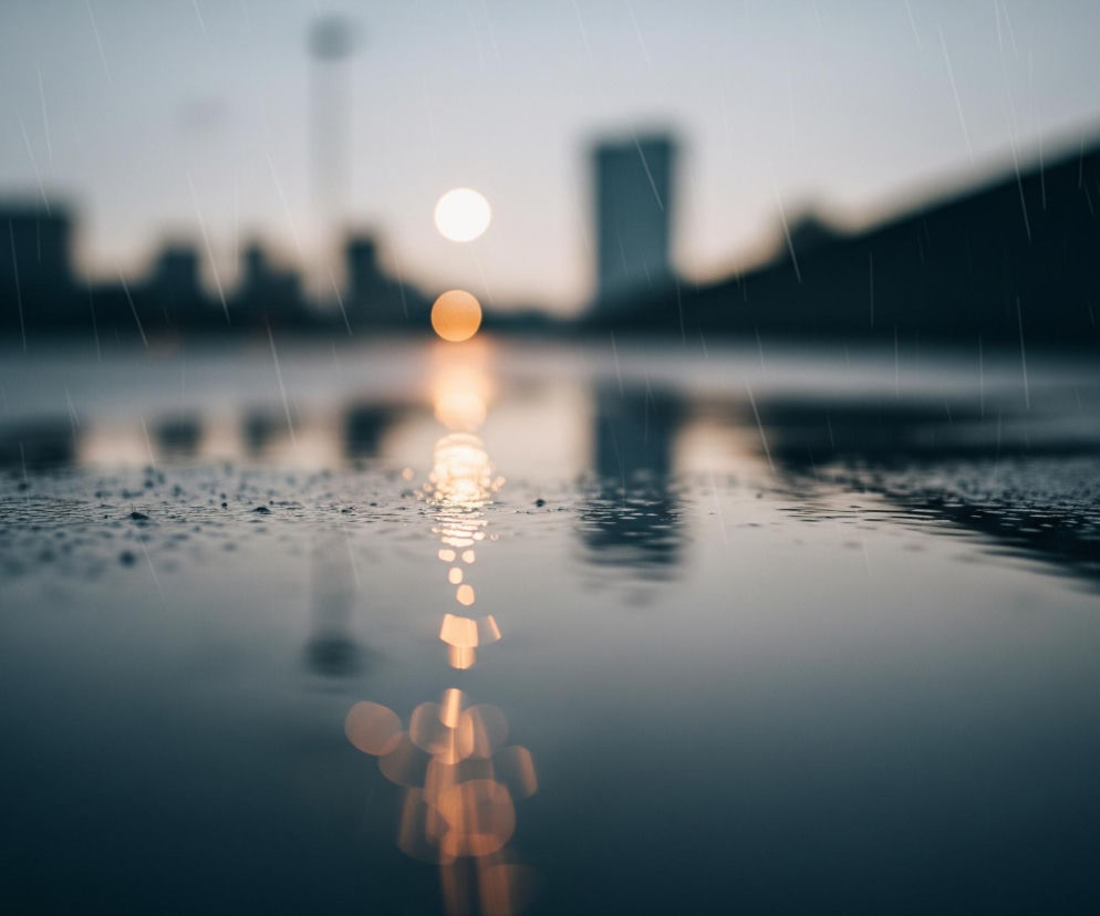 Powierzchnia wody, pokryta licznymi kroplami deszczu, zajmuje dolną połowę obrazu, odbijając smugę pomarańczowego światła biegnącą od środka w dół. W tle widoczna jest rozmyta panorama miasta z ciemnymi sylwetkami budynków, a pośrodku horyzontu świeci okrągłe, jasne słońce, którego blask odbija się na wodzie. Cały obraz jest przesiąknięty deszczem, widocznym jako ukośne, białe smugi na tle ciemnego nieba i rozmytych kształtów miejskiej zabudowy. Kolorystyka utrzymana jest w chłodnych odcieniach szarości, błękitu i ciemnego granatu, kontrastujących z ciepłym, pomarańczowym światłem.