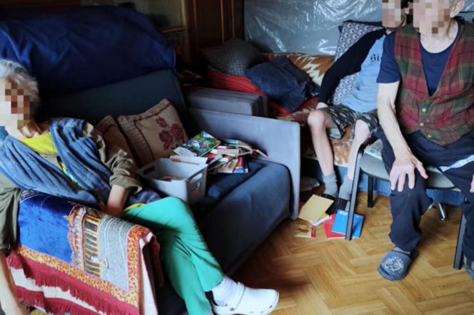 Trzy osoby siedzące w zagraconym pokoju. O historii więzionej przez 30 lat Magdaleny przeczytasz na Mediateka.