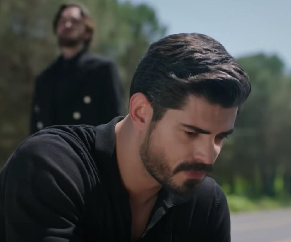 Cihan, główny bohater serialu Panna młoda, z pochyloną głową, ubrany w ciemną koszulkę polo, na drodze podczas napaści w 77. odcinku. W tle widoczny zamachowiec, który zlecił morderstwo Cihana. Więcej o dramatycznych losach bohaterów na Super Seriale.