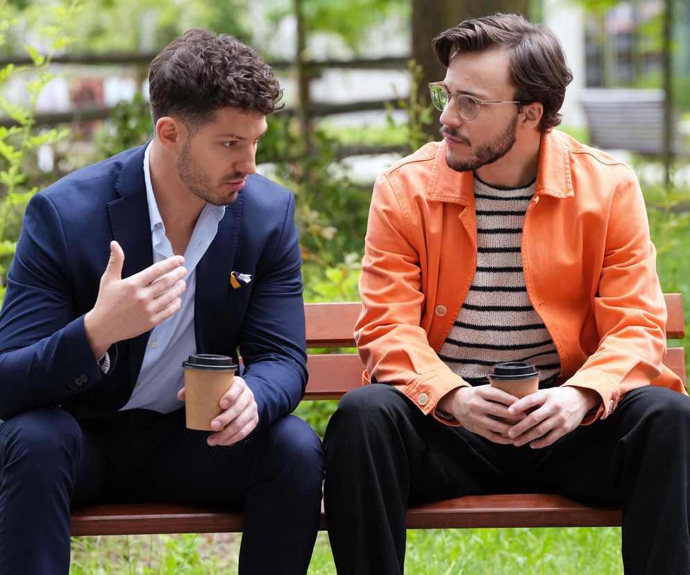 Aktorzy Jakub Dmochowski (Kajtek) w granatowym garniturze i Jasper Sołtysiewicz (Justin) w pomarańczowej kurtce i okularach siedzą na ławce w parku, rozmawiając i trzymając kubki kawy, co symbolizuje ich sojusz. Więcej o ich spisku w Barwach szczęścia przeczytasz na Super Seriale.