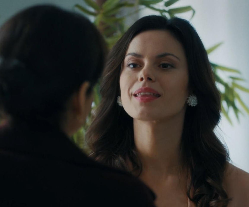 Kasia (Paulina Lasota) z serialu M jak miłość, ubrana w jasną suknię i ozdobne kolczyki, patrzy z lekkim uśmiechem na rozmazaną sylwetkę innej osoby, w tle widać roślinę. Więcej informacji o serialowych perypetiach bohaterów znajdziesz na Super Seriale.