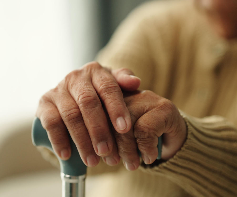 Pomarszczone dłonie starszej osoby o ciemnej karnacji, spoczywające na rączce laski lub kuli, symbolizującej wsparcie w poruszaniu się. Postać ubrana w beżowy sweter, a tło jest jasne i rozmyte, co podkreśla skupienie na dłoniach. Na portalu Poradnik Zdrowie znajdziesz ważne informacje o świadczeniach dla osób z chorobami przewlekłymi.