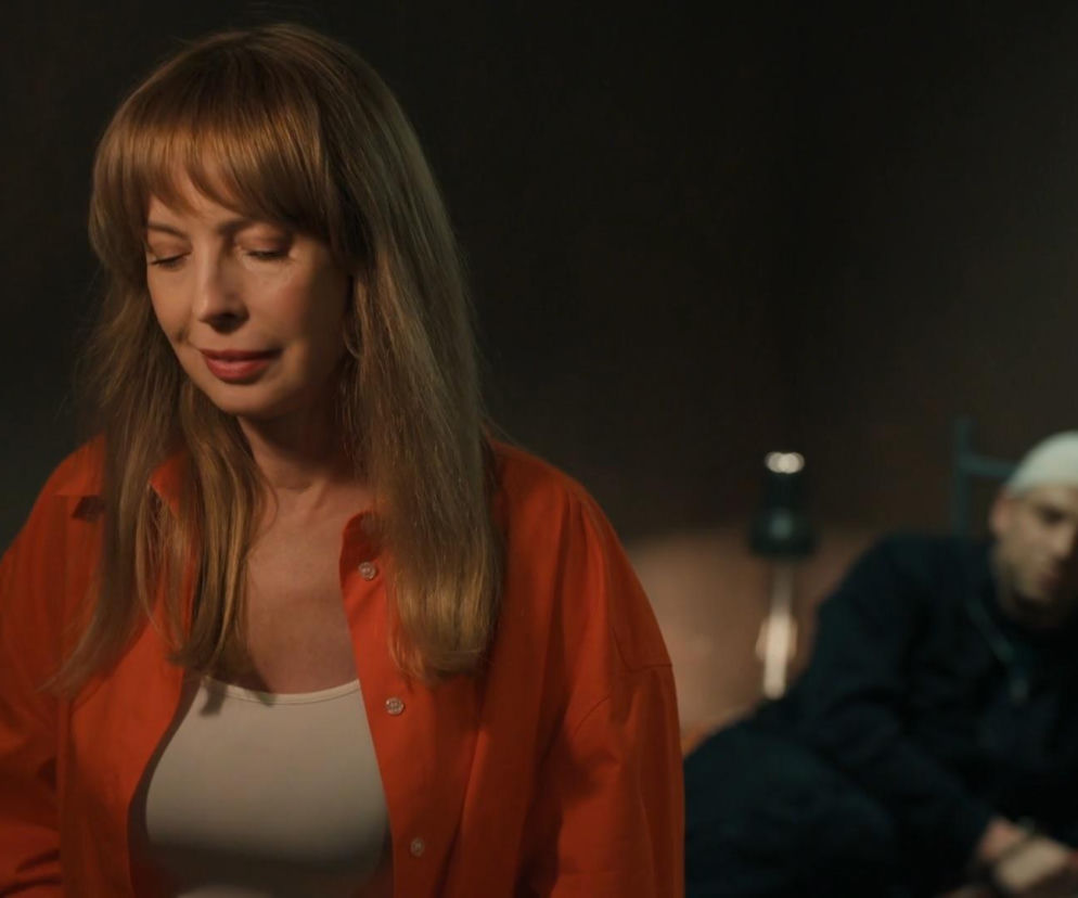 Aktorka Katarzyna Tlałka jako Agnieszka w pomarańczowej koszuli, z zamkniętymi oczami, z tyłu w tle siedzi aktor Michał Rolnicki jako Łukasz, więziony w odcinku serialu „Barwy szczęścia”. Więcej o losach bohaterów na Super Seriale.