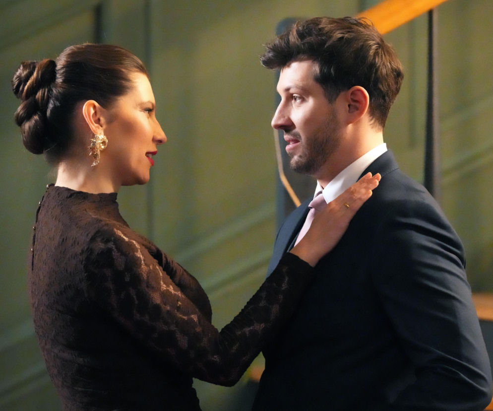 Karolina w czarnej koronkowej sukience, z upiętymi włosami i dużą złotą kolczykiem, kładzie dłoń na ramieniu Justina, który jest w garniturze z różowym krawatem, oboje patrzą na siebie z troską, widoczne schody i świece w tle. Więcej emocjonujących scen znajdziesz na stronie Super Seriale.