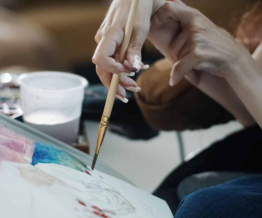 Dłoń z pędzlem malująca akwarelowy obraz na kartce, gdzie widać szkic sylwetki i kolorowe plamy. Scena symbolizuje nadzieję i nowe możliwości dla pacjentów ze SMA, podkreślając znaczenie powrotu do sprawności i samodzielności, o czym przeczytasz więcej na Poradnik Zdrowie.