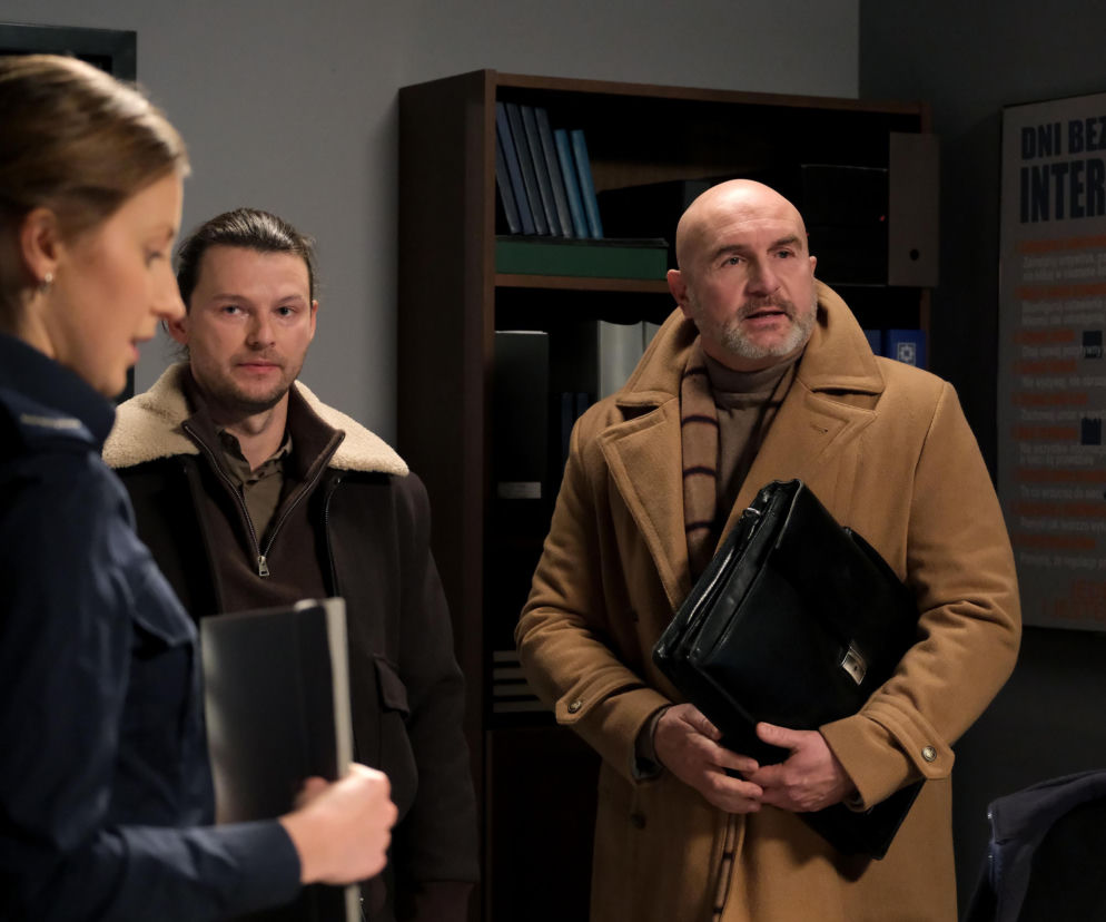 Natalka (Dominika Suchecka) w mundurze policjantki, z dokumentami, rozmawia z Mikołajem (Mateusz Kościukiewicz) i mężczyzną w beżowym płaszczu z teczką, w tle regał i plakat Dzień Bezpiecznego Internetu. Scena z serialu M jak miłość, o czym więcej na Super Seriale.