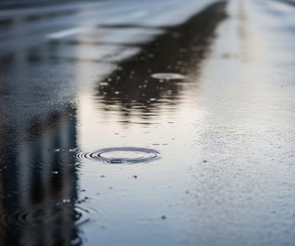 Kropla deszczu tworzy koncentryczne kręgi na kałuży pokrywającej ciemny asfalt ulicy. Po lewej stronie kałuży odbijają się zamazane, pionowe kształty budynków w odcieniach brązu i szarości. W tle, droga zwilżona deszczem rozciąga się w głąb, tworząc efekt rozmycia, a na jej powierzchni widać białe linie pasów ruchu.