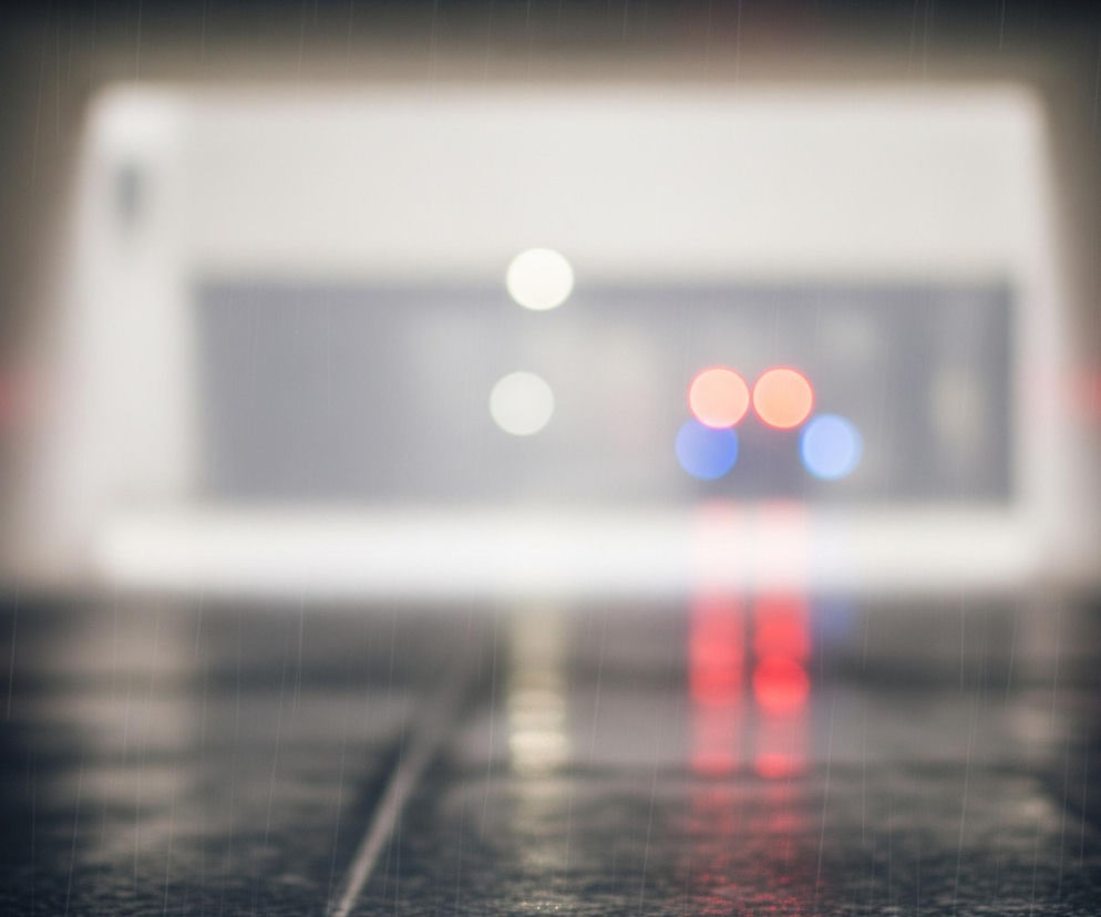 Ciemne otoczenie, z widocznymi licznymi, cienkimi, pionowymi liniami symulującymi padający deszcz. Centralnie, w oddali, znajduje się rozmyty, jasny, prostokątny kształt, przypominający wjazd do tunelu lub przejścia. W tle, wewnątrz tego kształtu, widoczne są rozmazane źródła światła: dwie białe kule na górze oraz dwie czerwone i dwie niebieskie kule poniżej, rozmieszczone symetrycznie. Czerwone światła odbijają się na mokrej, ciemnej powierzchni na dole obrazu.