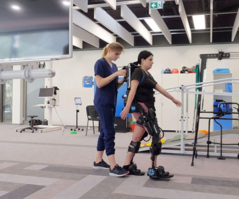 Kobieta w egzoszkielecie rehabilitacyjnym ćwiczy chód z pomocą fizjoterapeutki, symbolizując nadzieję w walce z ataksją Friedreicha. Nowoczesna terapia, o której pisze Poradnik Zdrowie, daje szansę na odzyskanie sprawności.