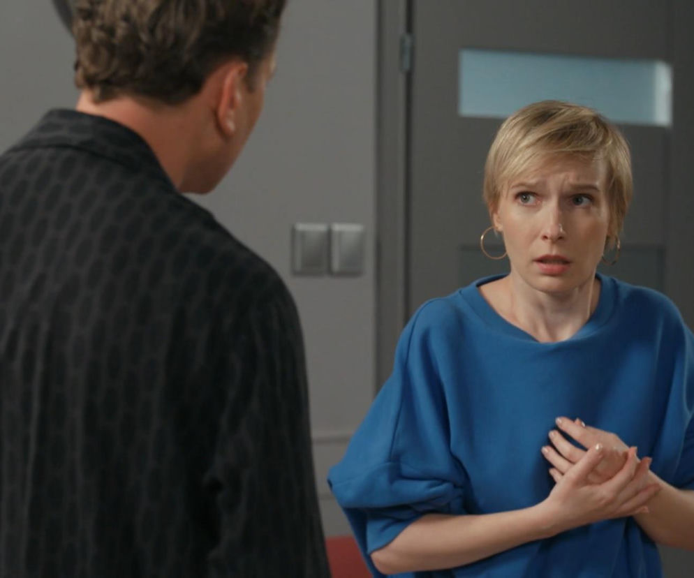 Kobieta o jasnych, krótkich włosach, Sonia (Weronika Nockowska), w niebieskiej bluzie, z przestrachem na twarzy, trzymająca dłonie przy klatce piersiowej, patrzy na stojącego tyłem mężczyznę, Roberta Stefaniaka (Paweł Ławrynowicz), w ciemnej koszuli z wzorami. Scena z serialu Barwy szczęścia, opisująca dramatyczne wydarzenia, które można znaleźć na stronie Super Seriale.