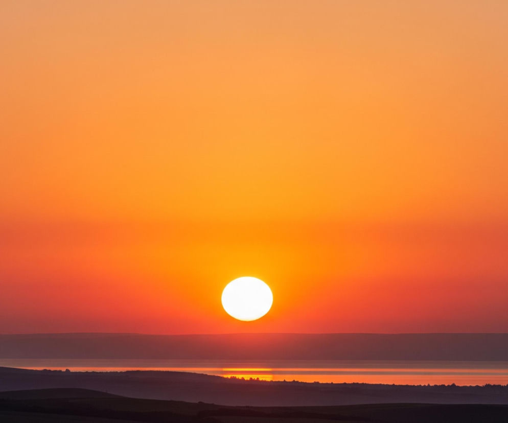 Na zdjęciu dominuje jaskrawe, pomarańczowo-czerwone niebo, przechodzące od jaśniejszych, żółto-pomarańczowych odcieni u góry do głębokiej czerwieni bliżej horyzontu. Centralnie umiejscowione jest jasne, białe słońce, odbijające swoje światło na powierzchni wody, tworząc poziomy pas jasności. Poniżej horyzontu rozciąga się ciemny krajobraz z widocznymi zarysami wzgórz i pasów zieleni, co kontrastuje z intensywnymi barwami nieba.