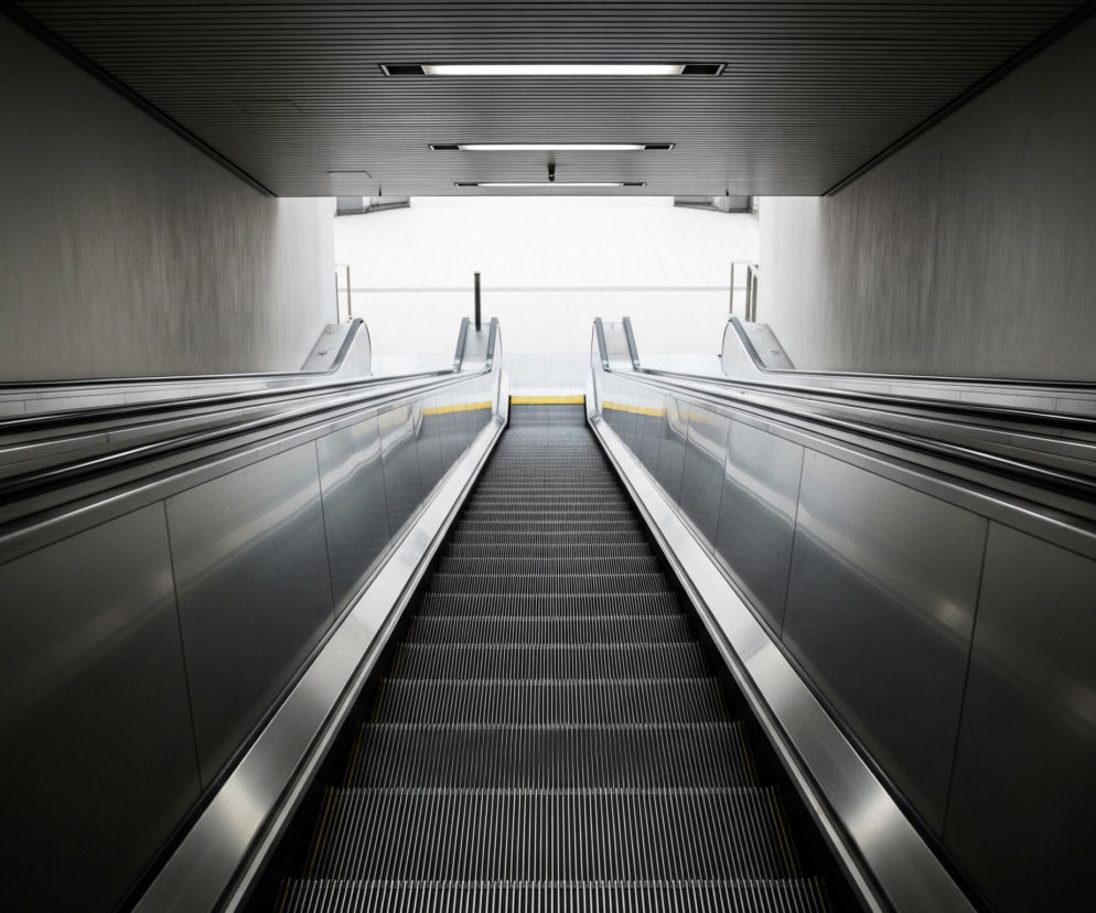 Długie, proste schody ruchome prowadzą w górę, kończąc się w jasnym, białym otwartym przejściu. Stopnie schodów są ciemnoszare z cienkimi, metalicznymi liniami, a po ich bokach widać szerokie, błyszczące, srebrne balustrady z czarnymi poręczami. Ściany boczne tunelu, w którym znajdują się schody, są jasnoszare, podczas gdy sufit w górnej części jest ciemniejszy, metalowy, z widocznymi prostokątnymi lampami.