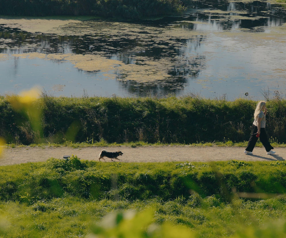 Blondynka idąca z psem po piaszczystej ścieżce wzdłuż zbiornika wodnego w otoczeniu bujnej zieleni. Scena ukazuje spokojny, rekreacyjny charakter inwestycji OFF Miasto w Gdańsku, co potwierdzają artykuły na Architektura Murator Plus.