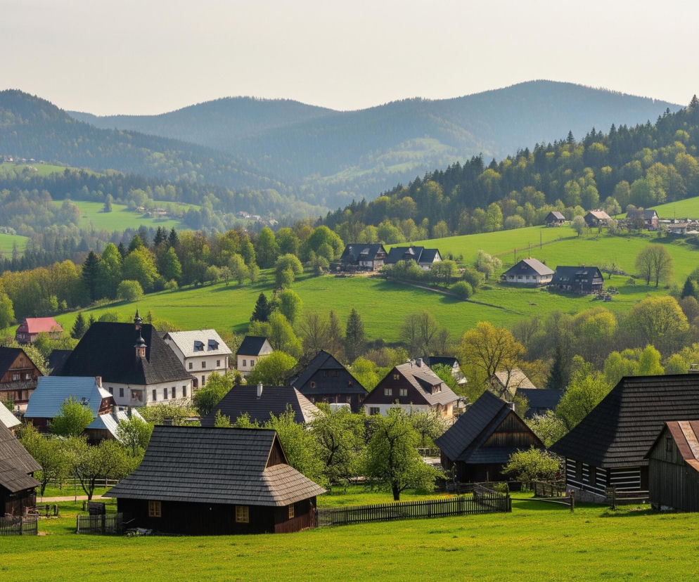 Rozległy krajobraz przedstawia malowniczą wioskę położoną w dolinie, otoczoną zielonymi wzgórzami i zalesionymi zboczami. Na pierwszym planie dominują ciemne, drewniane domy z tradycyjnymi, spadzistymi dachami pokrytymi gontem, oddzielone od siebie niskimi, drewnianymi płotami i sadami z kwitnącymi drzewami. W tle widać więcej domów o różnorodnej architekturze, od jasnych, tynkowanych budynków z ciemnymi dachami po domy o drewnianych fasadach, rozrzucone na falujących, intensywnie zielonych łąkach. Dalszy plan to pasma wzgórz porośniętych gęstym lasem, których barwy bledną i stają się bardziej mgliste w miarę oddalania się w kierunku jasnego, lekko zamglonego nieba.