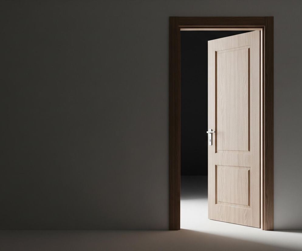 Po prawej stronie obrazu widać drewniane drzwi z klamką, lekko uchylone, z wyraźnym, ciemnym obramowaniem. Za drzwiami znajduje się ciemna przestrzeń, kontrastująca z jasnym światłem padającym na podłogę przed nimi, tworzącym rozmyty cień. Ściana po lewej stronie jest ciemnoszara, a podłoga jest jasnoszara, delikatnie oświetlona.