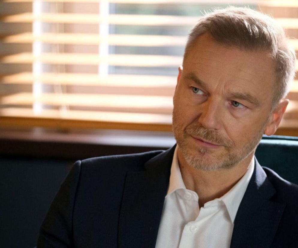 Krystian Wieczorek jako Andrzej Budzyński w eleganckiej granatowej marynarce i białej koszuli, patrzący w bok z zamyślonym wyrazem twarzy, na tle okna z żaluzjami. Aktor wciela się w postać prawnika, którego życie prywatne zostanie wystawione na próbę przez nową klientkę Annę, o czym przeczytasz na Super Seriale.