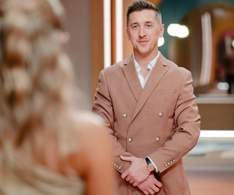 Kamil z Love Is Blind w brązowej dwurzędowej marynarce i białej koszuli, z dłońmi splecionymi na wysokości pasa, uśmiecha się w stronę niewyraźnej blondynki tyłem. Tło stanowi studio z jasnymi ścianami i łukowatym przejściem, gdzie można przeczytać więcej o nim na Super Seriale.