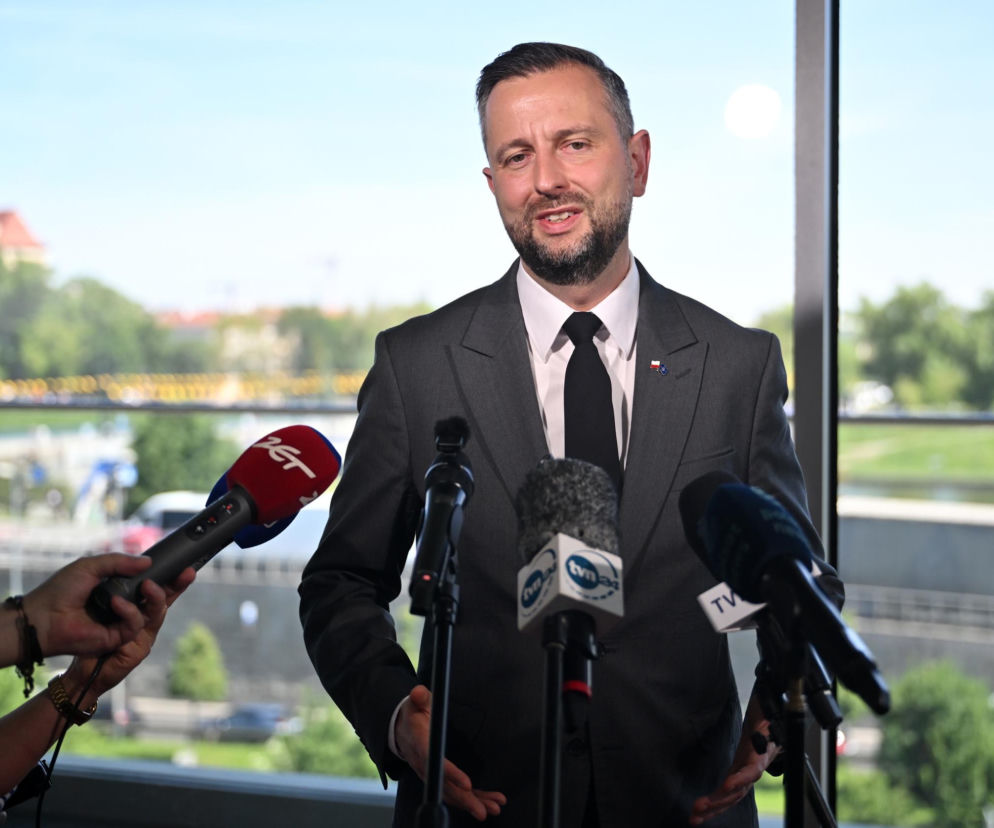 Władysław Kosiniak-Kamysz w szarym garniturze, białej koszuli i czarnym krawacie, z przypinką flagi Polski na marynarce, podczas konferencji prasowej. W tle zieleń i fragmenty zabudowań. O majątku polityka przeczytasz na portalu Super Biznes.
