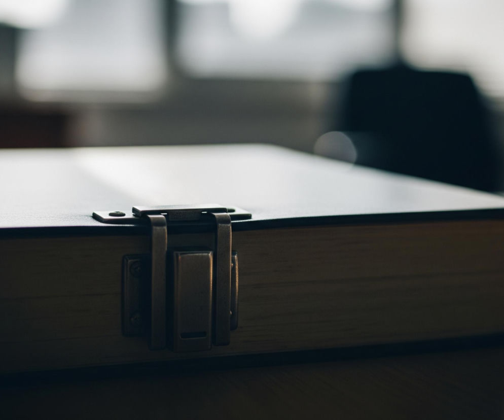 Duża, grubo oprawiona księga z metalowym zapięciem leży na drewnianym blacie, zajmując większą część kadru. Zapięcie, wykonane z metalu w kolorze brązu lub miedzi, znajduje się na środku frontu księgi. Brzegi kartek, w odcieniach beżu i jasnego brązu, są widoczne po prawej stronie, oświetlone promieniami światła, tworzącymi odblaski na powierzchni blatu. Tło jest rozmyte, ukazując jasne, niewyraźne kształty, sugerujące okna i ciemny, zarysowany obiekt, przypominający oparcie krzesła.