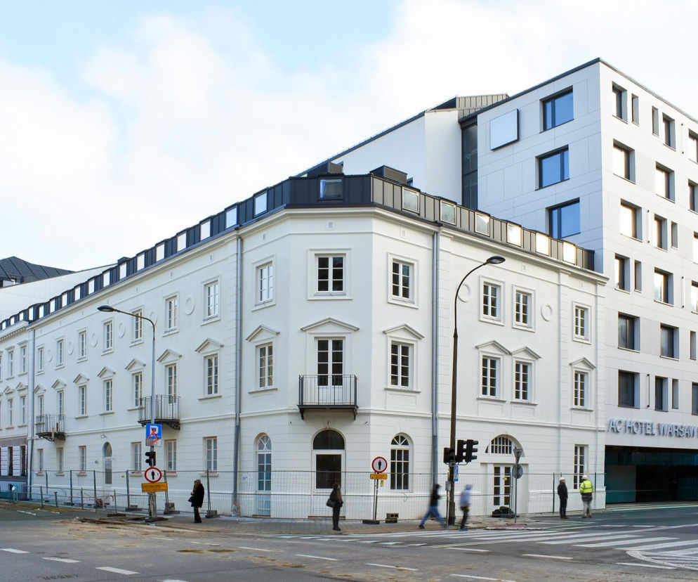 Odrestaurowana, biała kamienica Karola Mintera na warszawskiej Pradze-Południe, z secesyjną elewacją i balkonami, połączona z nowoczesną, jasną bryłą hotelu AC Hotel Warsaw Port Praski, widoczna z poziomu ulicy z przechodniami i barierkami remontowymi. Więcej o renowacji kamienic przeczytasz na Architektura Murator Plus.