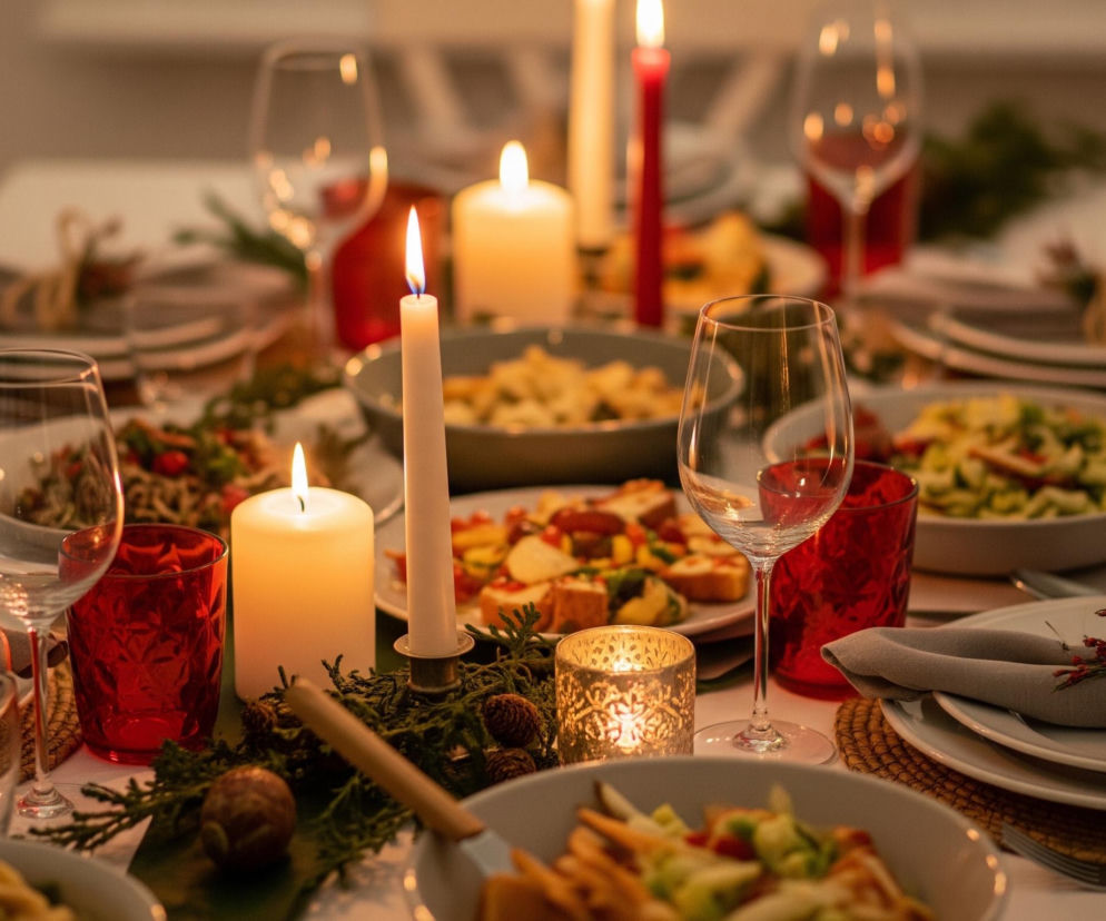 Stół jest nakryty do uroczystego posiłku i oświetlony blaskiem świec. Na białym obrusie ułożono liczne naczynia, takie jak miski z jedzeniem, talerze z serwetkami przewiązanymi sznurkiem i ozdobionymi gałązkami. Pomiędzy zastawą znajdują się kieliszki do wina oraz czerwone szklanki, a także świece o różnej wysokości – niektóre białe, inne czerwone – z palącymi się płomieniami. Elementy dekoracyjne stołu to zielone gałązki, szyszki i świeczniki z ażurowym wzorem.