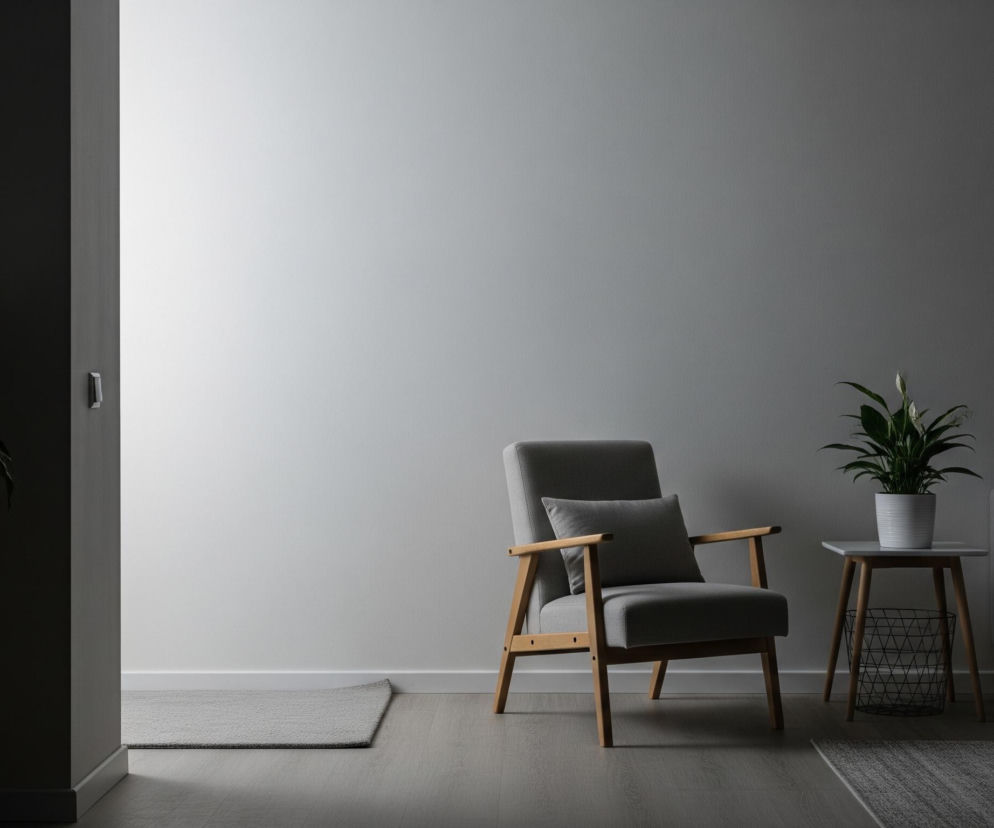 Jasno oświetlony fragment białej ściany zajmuje centralną część kadru, przechodząc w ciemniejsze odcienie szarości po prawej stronie. Pośrodku znajduje się szary fotel z drewnianymi podłokietnikami i nóżkami, a obok niego, po lewej, niewielki, jasny dywanik na drewnianej podłodze. Po prawej stronie fotela stoi okrągły, biały stolik z doniczkową rośliną o zielonych liściach i białych kwiatach, obok którego widoczny jest fragment ciemnej sofy i drewniany stolik kawowy. W lewej części obrazu, w ciemnym korytarzu, widoczna jest lampa podłogowa z szerokim, szarym abażurem oraz fragment innej rośliny w doniczce, natomiast w górnym prawym rogu na ciemnej ścianie wisi czarna ramka z obrazem przedstawiającym liście.