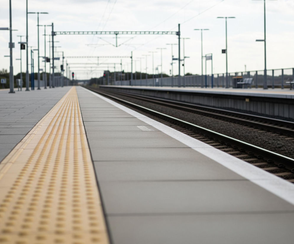 Na pierwszym planie, z lewej strony, widoczny jest szary peron kolejowy, wyłożony prostokątnymi płytami. Wzdłuż krawędzi peronu biegnie jasnobrązowy, fakturowany pas ostrzegawczy z regularnie rozmieszczonymi wypustkami. Po prawej stronie peronu znajduje się torowisko z dwoma rzędami stalowych szyn ułożonych na ciemnym tłuczniu i drewnianych podkładach. W oddali, wzdłuż torowiska, ciągnie się rząd słupów trakcyjnych i oświetleniowych, oraz widoczne są fragmenty drugiego peronu z barierkami i ławkami. Całość pokrywa jasne, zachmurzone niebo.
