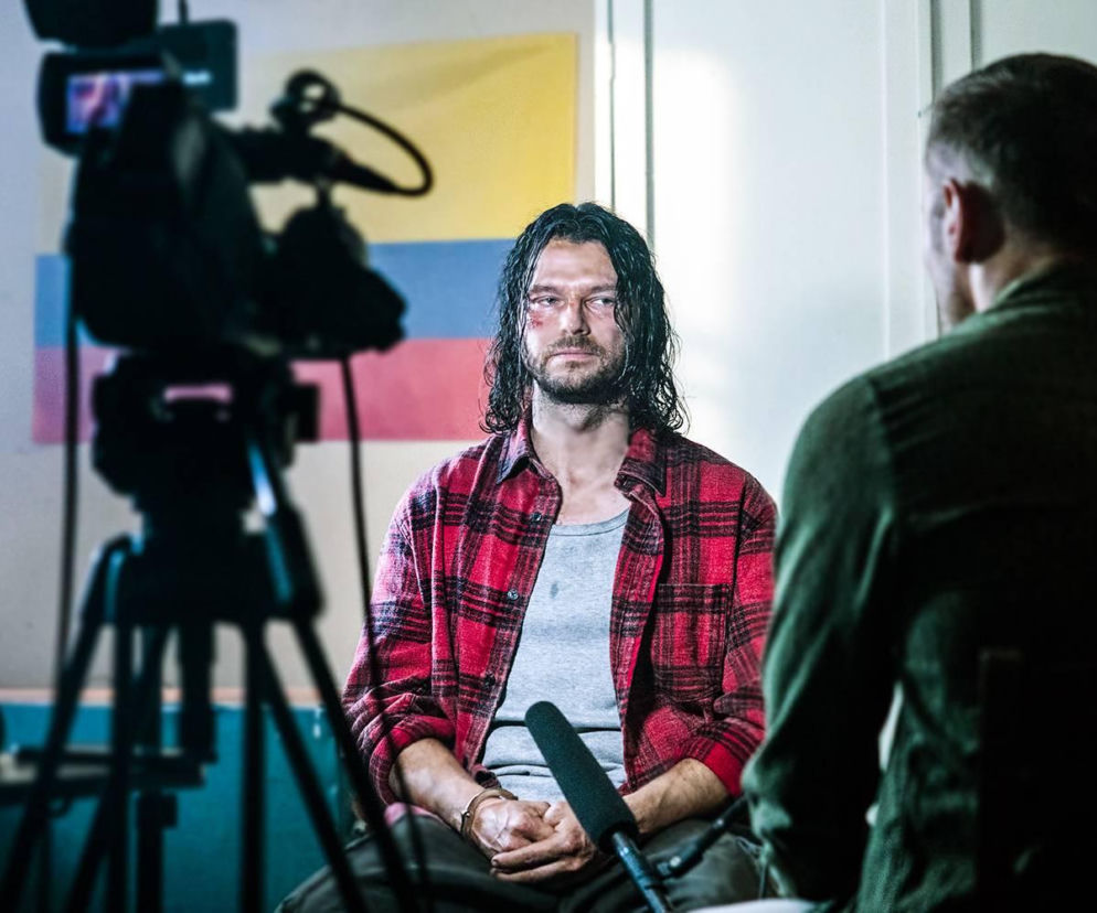 Mężczyzna z długimi, ciemnymi włosami i zarostem, ubrany w czerwoną koszulę w kratę, siedzi, mając na nadgarstkach kajdanki. Na jego twarzy widać otarcia. Obok leży mikrofon, a z boku widoczna jest kamera nagrywająca wywiad. Flaga Kolumbii w tle. Więcej na ten temat znajdziesz na Super Seriale.