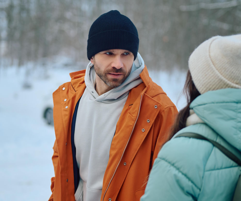 Mężczyzna w czarnej czapce i pomarańczowej kurtce patrzy na rozmówczynię w zimowym otoczeniu. Zdjęcie ilustruje dynamiczne relacje międzyludzkie, nawiązujące do wątków serialu Na Wspólnej, o którym przeczytasz więcej na Super Seriale.
