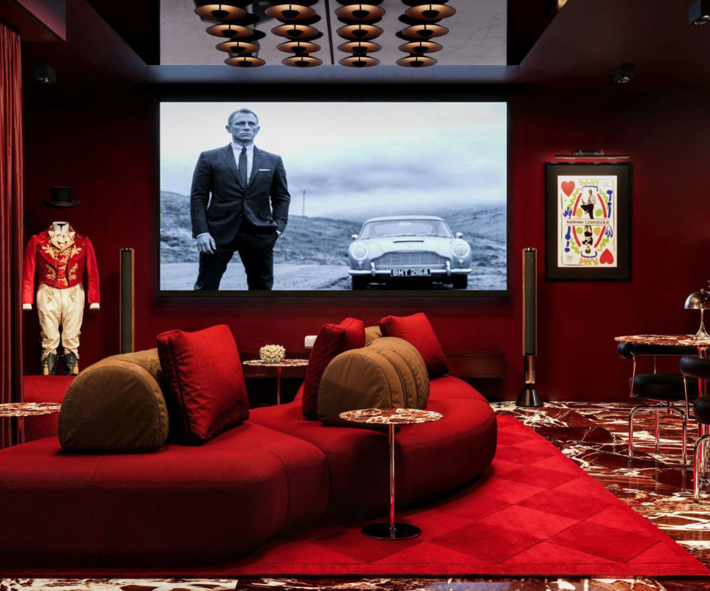 Wnętrze apartamentu kolekcjonerskiego Kino Archicom Collection, inspirowanego studyjnymi salami filmowymi, z czerwonymi sofami, aksamitnymi zasłonami i dużym ekranem. Na ekranie widać scenę z Jamesem Bondem i samochodem Aston Martin, obok stoi oryginalny kostium Johnny'ego Deppa z filmu „Marzyciel” oraz plakat Wojciecha Fangora do „Niewinnych czarodziejów”. Więcej o luksusowych inwestycjach przeczytasz na portalu Architektura Murator Plus.