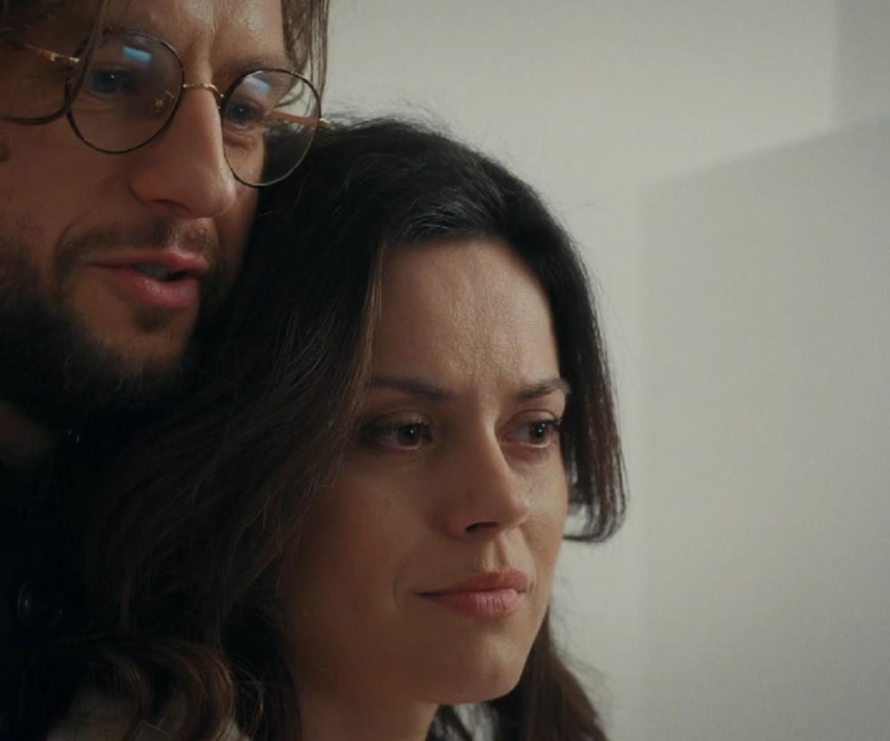 Zbliżenie na Kasię (Paulina Lasota) i Mariusza (Mateusz Mosiewicz) z serialu M jak miłość. Kasia z zasmuconą miną patrzy przed siebie, podczas gdy Mariusz, obejmując ją od tyłu, sprawia wrażenie zadowolonego. Zdjęcie ilustruje scenę, gdzie Kasia udaje szczęście, pomimo wewnętrznych wyrzutów sumienia, o czym możesz przeczytać na Super Seriale.