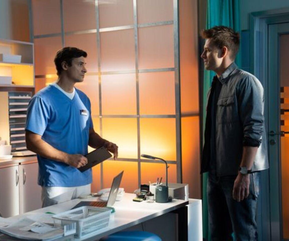 Lekarz Krzysztof Radwan w niebieskim stroju medycznym i Mario w ciemnej kurtce stoją naprzeciwko siebie w gabinecie. Rozmawiają o pojednaniu i ratowaniu życia siostry Mario, co wywołało zaskakujące wydarzenia w serialu Na dobre i na złe. Więcej na temat tej intrygującej historii znajdziesz na Super Seriale.