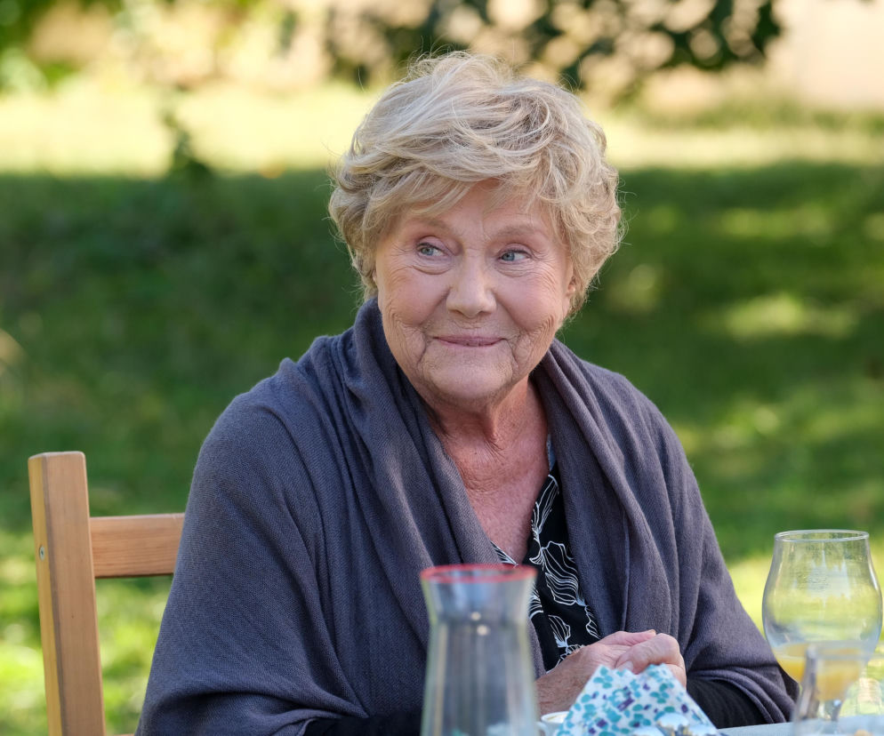 Uśmiechnięta Barbara (Teresa Lipowska) w szarym swetrze i bluzce w biało-czarne wzory siedzi przy stole w ogrodzie. Na stole widać dzbanek i szklanki. W tle rozmazana zieleń. Mostowiakowa odzyskuje humor w M jak miłość. Więcej o serialu przeczytasz na stronie Super Seriale.