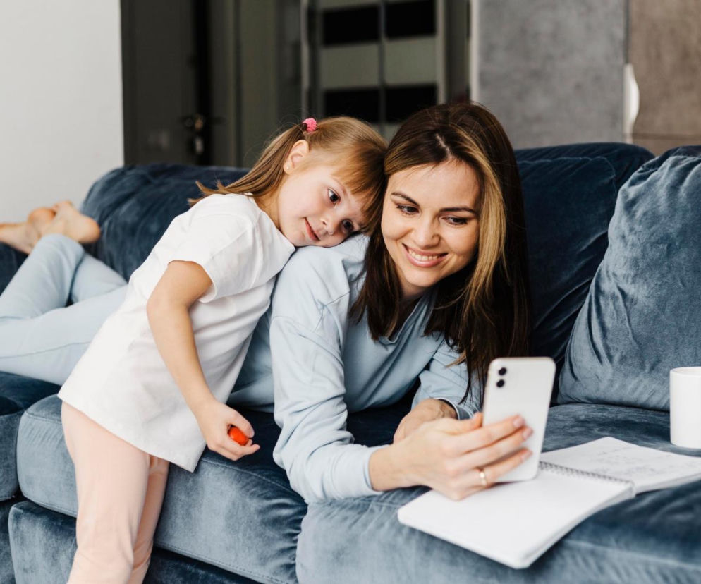 Uśmiechnięta mama z córką patrzą w telefon na kanapie. O stresie przez aplikacje przeczytasz na SE.