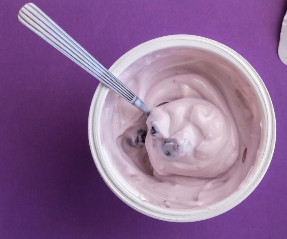 Kubek jogurtu z fioletową zawartością i łyżeczką zanurzoną w środku, leżący na fioletowym tle. Obok widoczne wieczko. Obraz odnosi się do ostrzeżenia GIS o szkle w jogurcie Lublanka, o czym przeczytasz na Super Biznes.