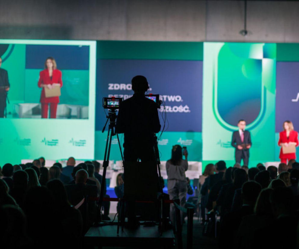 Sylwetka kamerzysty na tle sceny z prelegentami oraz napisem ZDROWE PAŃSTWO. ZDROWIE. PRZYSZŁOŚĆ. podczas XI Kongresu Wyzwań Zdrowotnych w Katowicach, gdzie dyskutowano o nowych strategiach dla zdrowia. Więcej na temat wydarzenia przeczytasz na Poradnik Zdrowie.