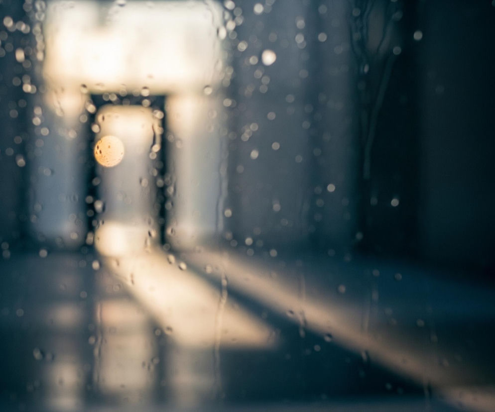Przedni plan to szyba pokryta licznymi, rozproszonymi kroplami wody o nieregularnych kształtach i rozmiarach, tworzącymi efekt bokeh. Rozmyte tło ukazuje ciemny korytarz lub przejście, z wyraźnym, jasnym prostokątnym otworem w głębi, prawdopodobnie oknem lub wejściem, z którego wpada skośny promień światła. Światło to tworzy podłużny, jasny kształt na podłodze, a w jego obszarze widoczna jest duża, okrągła, złocista plama świetlna, będąca zapewne odblaskiem. Dominują odcienie granatu, szarości i brązu, z ciepłymi, jasnymi akcentami światła.