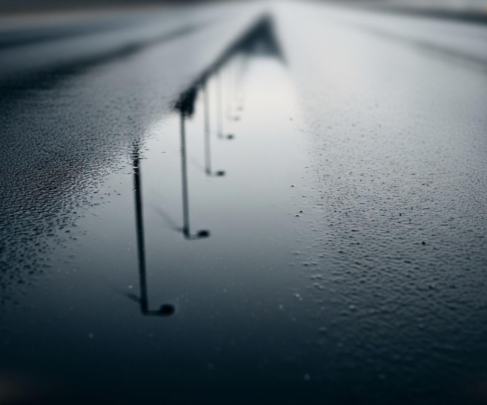 Na pierwszym planie widoczna jest mokra, ciemna nawierzchnia pokryta licznymi kroplami wody, które tworzą nieregularne wzory. Od lewej strony obrazu, wzdłuż jego osi, rozciąga się rzutowanie odbitego w wodzie ciągu pionowych obiektów, z których niektóre są zakończone niewielkimi, zaokrąglonymi elementami. Odbicia te stają się coraz bardziej rozmyte i niewyraźne w miarę oddalania się w głąb obrazu, gdzie ciemniejsza powierzchnia przechodzi w jaśniejszy, zamglony horyzont. W dolnej części, po lewej stronie, oraz w górnym prawym rogu, widoczne są subtelne żółtawe refleksy światła.