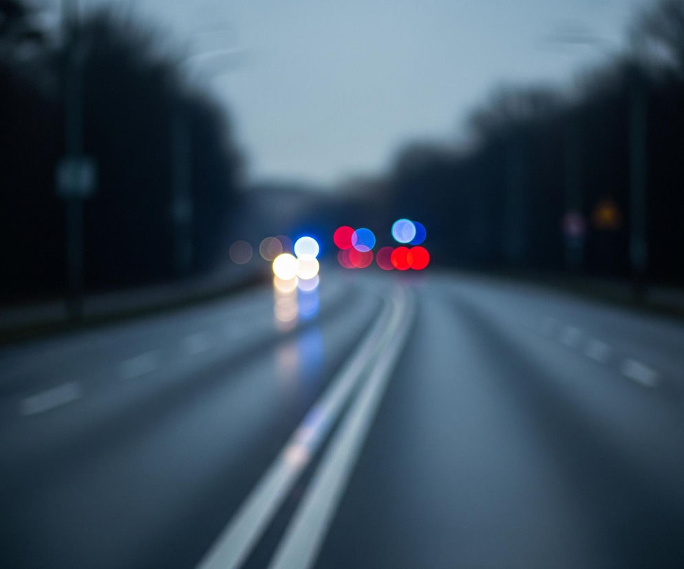 Asfaltowa droga, oznaczona podwójną ciągłą białą linią na środku oraz przerywanymi białymi liniami po bokach, rozciąga się w głąb obrazu, stopniowo zwężając się i znikając w nieostrym tle. Po obu stronach jezdni rosną ciemne, niewyraźne drzewa, tworząc rodzaj tunelu. Na drodze, w oddali, widać rozmazane światła pojazdów, głównie czerwone, niebieskie i żółte, z ich odbiciami widocznymi na mokrej nawierzchni. Górna część obrazu to jasnoszare niebo, przechodzące w ciemniejsze tony ku górze.