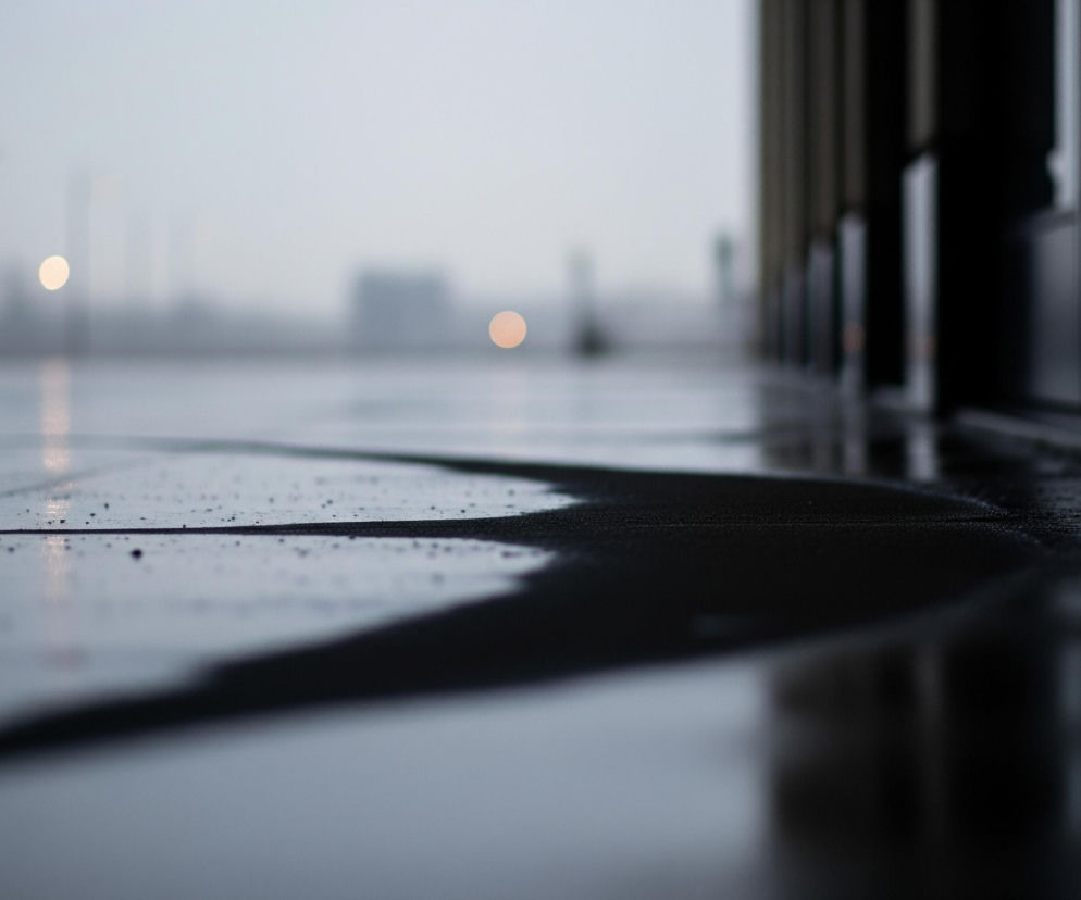 Mokrą, ciemną nawierzchnię z widocznymi kroplami wody, która odbija otoczenie. Po prawej stronie kadru znajduje się ciemna ściana budynku lub konstrukcji, tworząca pionowe pasy, z widocznymi drobnymi teksturami. W oddali, na szarym, rozmytym tle, znajdują się rozświetlone, okrągłe obiekty, prawdopodobnie światła uliczne, oraz niewyraźne zarysy innych budynków lub słupów.
